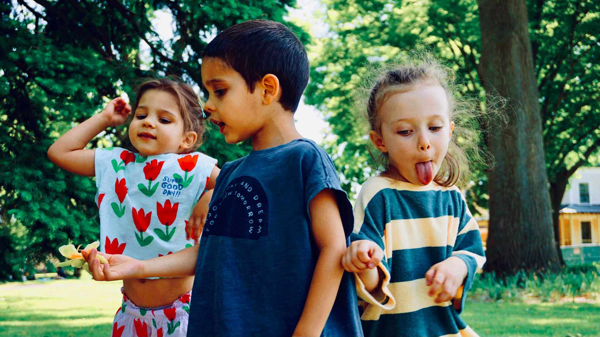 Three children standing in a park, smiling and playing with nature.